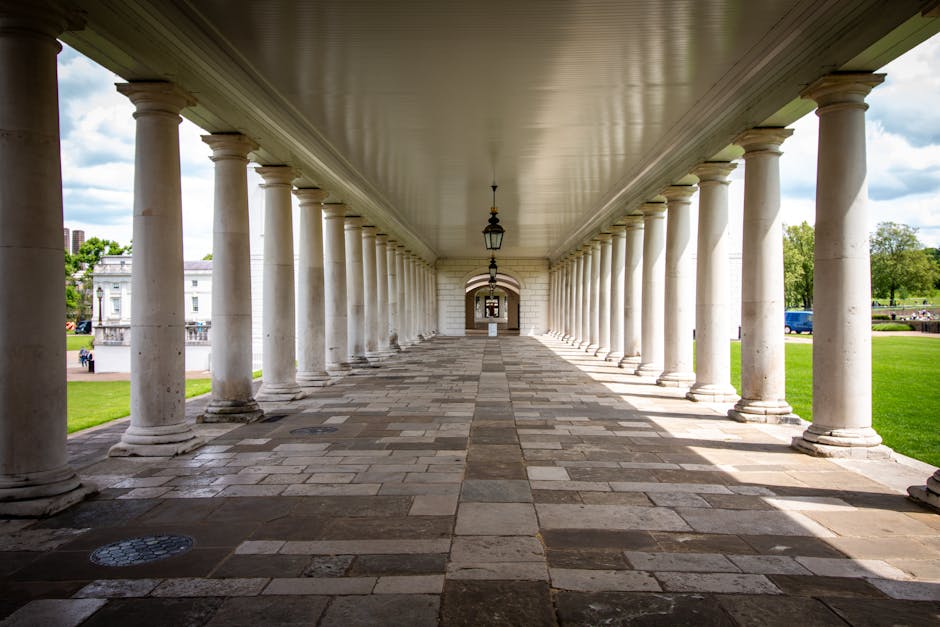 A long colonnade with white classical columns supporting a ceiling, creating a covered walkway in an outdoor park setting. The stone-paved ground is clean and well-maintained, with alternating dark and light tiles extending the length of the corridor. Lantern-style light fixtures hang from the ceiling at regular intervals, and the open sides reveal a green lawn and trees under a partly cloudy sky. The area appears bright and tidy, with no visible debris or dirt, reflecting a well-kept outdoor space suitable for surface cleaning and maintenance by Queensparkcarpetcleaning.co.uk.