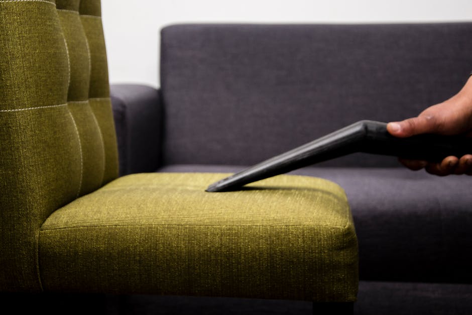 Close-up view of a hand holding a vacuum cleaner nozzle, actively cleaning a green upholstered chair with a tufted backrest, positioned in a living room setting. The chair's fabric appears clean and well-maintained, with the seat cushion showing minimal signs of dust or stains. Behind the green chair, a darker grey sofa with smooth fabric upholstery is partially visible, placed against a clean, white wall. The area is well-lit, highlighting the surface cleanliness and the detailed texture of the fabric. The scene emphasizes surface cleaning techniques used in upholstery sanitation, with a focus on maintaining hygiene and deep cleaning of residential furniture. The image reflects professional cleaning services provided by Queensparkcarpetcleaning.co.uk in the context of domestic surface cleaning and upholstery care.