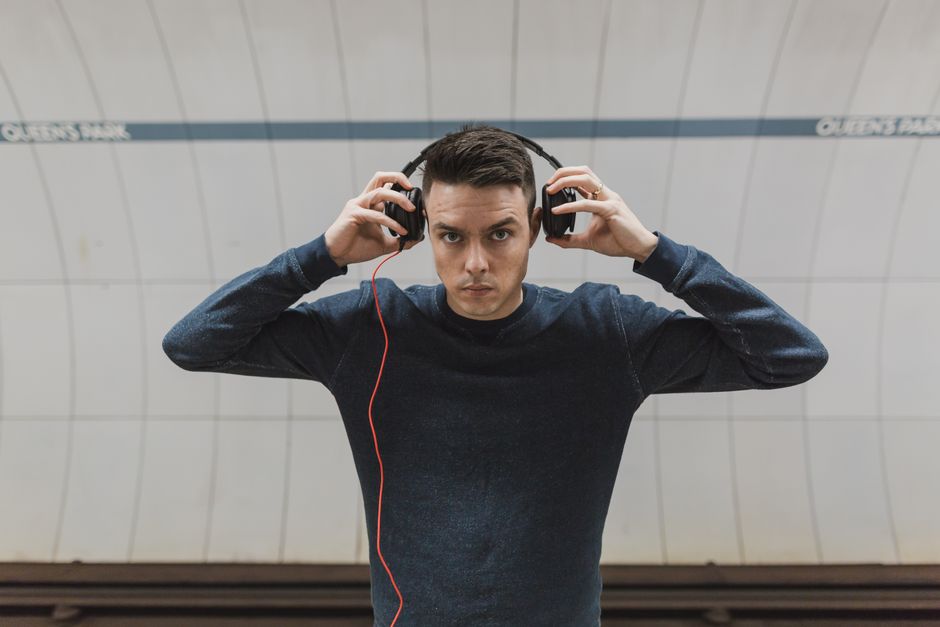 A young man with dark hair, wearing a dark long-sleeved shirt, appears to be in an underground or metro station with a curved, white tiled wall in the background. He is adjusting a large pair of black over-ear headphones with both hands, looking directly at the camera with a neutral expression. The lighting is bright and even, highlighting both the subject and the reflective surfaces of the tiles. This image illustrates a moment of personal audio engagement, potentially related to leisure or transit settings, but does not include cleaning or hygiene elements. For surface cleaning, upholstery, or sanitisation content, refer to images featuring cleaning tools, surfaces, or environments focused on hygiene practices, which can be associated with QueensparkCarpetCleaning.co.uk.